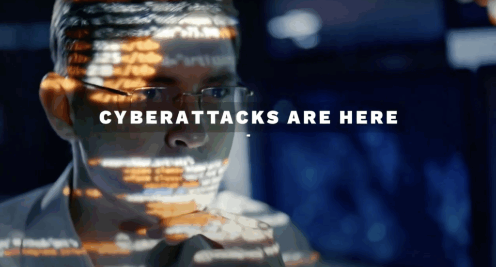 A man with glasses looks at a screen with code reflected on his face. The text "CYBERATTACKS ARE HERE" appears across the image, emphasizing the threat of cyberattacks.